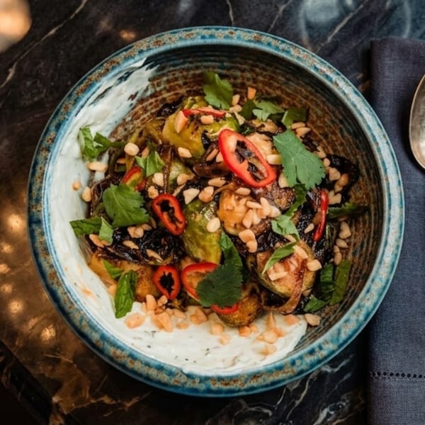 A bowl of roasted Brussels sprouts with red peppers, peanuts, herbs, and a creamy white sauce.
