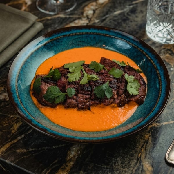 Grilled meat slices in a vibrant orange sauce, garnished with fresh cilantro in a blue bowl.
