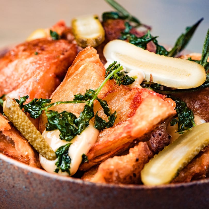 Close-up of crispy fried potatoes with creamy sauce, fresh herbs, and sliced pickles in a rustic bowl.