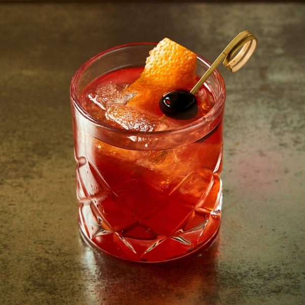 A vibrant red cocktail with ice, an orange peel, and a dark cherry on a skewer.