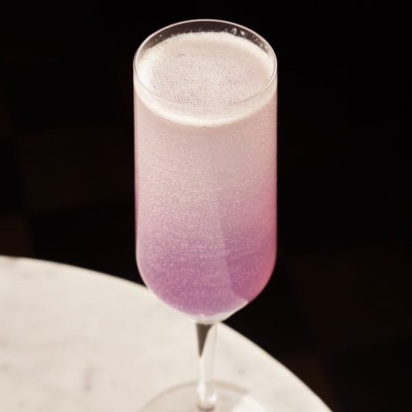 A tall, slender glass filled with a sparkling, gradient pink and purple cocktail on a white table.