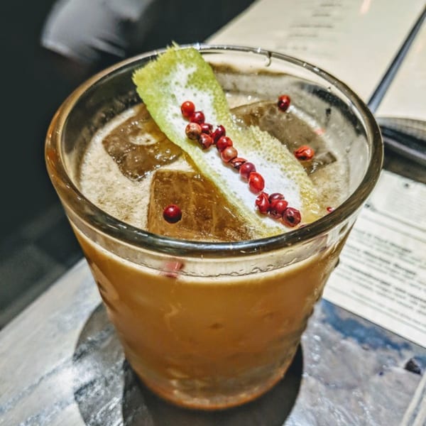 Close-up of a cocktail with ice, garnished with a lime peel and red peppercorns.