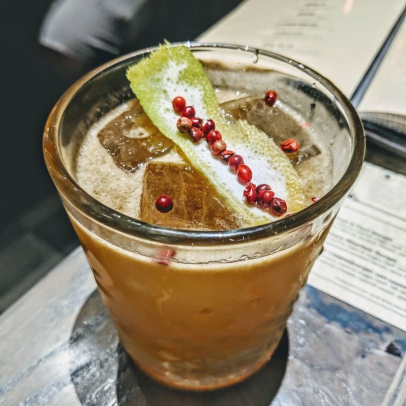 Close-up of a cocktail with ice, garnished with a lime peel and red peppercorns.